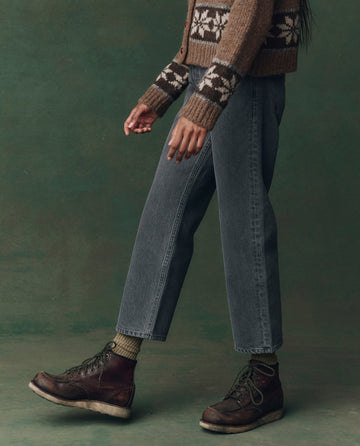 A person wearing a brown patterned sweater, THE GREAT. F25 D2 The Wayne Jean in Graphite Wash with Clean Hem, green socks, and brown lace-up boots stands against a green background.