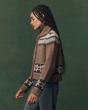 Person with long braids wears THE GREAT. F25 D2 The Nordic Scholar Cardigan in Hickory Fair Isle—an oversized brown knit with white and black geometric patterns—and blue jeans, standing against a green background.