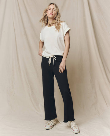 A person stands against a beige backdrop, donning The Boxy Crew tee in Washed White by THE GREAT. CORE KNITS, paired with black drawstring pants and crisp white sneakers.
