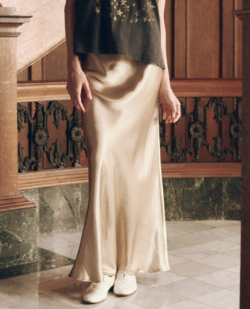 A person wearing a gold shimmer The Elastic Bias Skirt in a floor-length, elastic bias fit with white shoes.