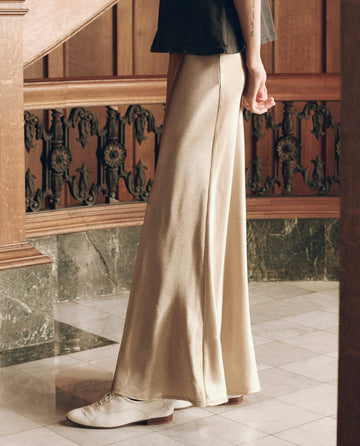The Elastic Bias Skirt in Gold Shimmer is a floor-length, gold-toned elastic bias skirt with a metallic finish, worn with a black t-shirt and white shoes.