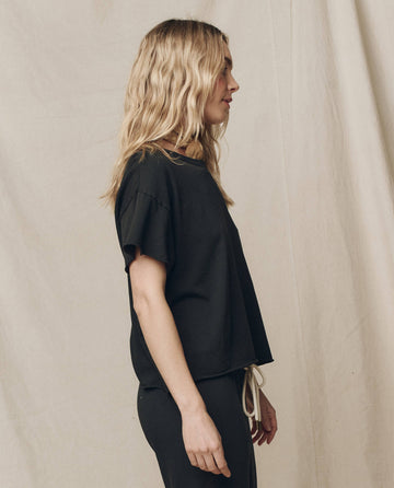 A wavy-haired blonde woman stands in profile wearing THE GREAT. CORE KNITS’ The Crop Tee in "Solid -- Almost Black," paired with matching pants, against a neutral fabric backdrop.