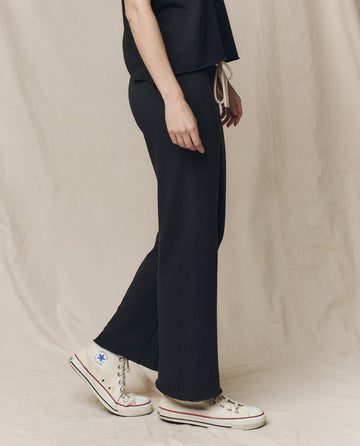 A person wearing THE GREAT. CORE KNITS' The Wide Leg Cropped Sweatpant in Almost Black, highlighted by a drawstring waist, is effortlessly paired with white high-top sneakers against a beige fabric background.