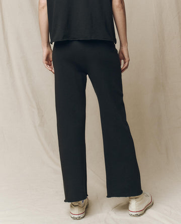 A person in "The Wide Leg Cropped Sweatpant" by THE GREAT. CORE KNITS, in Almost Black, and a matching top stands with their back to the camera, highlighting their white sneakers.