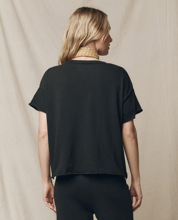 A person with long hair stands back to the camera, wearing The Crop Tee in Almost Black from THE GREAT. CORE KNITS, complemented by a light scarf, against a plain beige fabric backdrop. The ensemble subtly hints at comfort akin to super soft cotton.