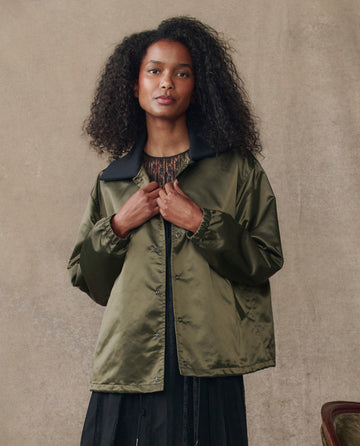 A woman with curly hair models THE GREAT. H25 COLLECTION’s The Satin Coach's Jacket in Bright Army over a patterned top and black skirt, standing before a beige textured background.