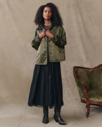 A woman stands indoors wearing THE GREAT. H25 COLLECTION The Satin Coach's Jacket -- Bright Army over a long black dress and black boots, next to a green upholstered wooden chair against a neutral backdrop.