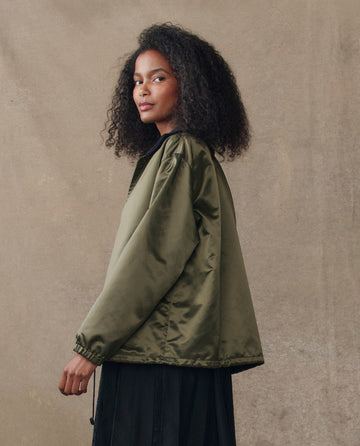 A woman with curly hair wears THE GREAT. H25 COLLECTION's The Satin Coach's Jacket in Bright Army over a black skirt, standing before a beige background and glancing over her shoulder.