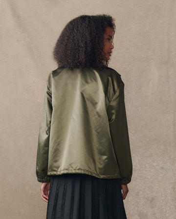 A person with curly hair wears THE GREAT. H25 COLLECTION's The Satin Coach's Jacket in Bright Army and a black skirt, standing with their back to the camera against a neutral background.