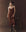 A person in THE GREAT. F25 D1’s The Legacy Dress -- Chocolate, with boots and a cowboy hat, stands beside a wooden stool holding gloves against a dark brown background.