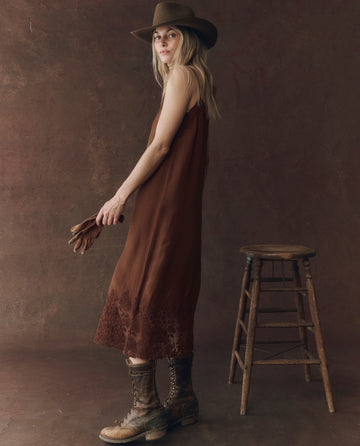 A woman stands by a wooden stool, wearing THE GREAT. F25 D1 The Legacy Dress in Chocolate with a hat, lace-up boots, and holding brown gloves against a textured brown backdrop.