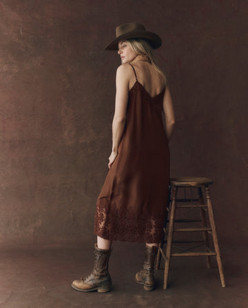 A woman in THE GREAT. F25 D1 The Legacy Dress – Chocolate and boots, wearing a wide-brim hat, stands by a wooden stool against a plain brown background, facing away while looking over her shoulder.