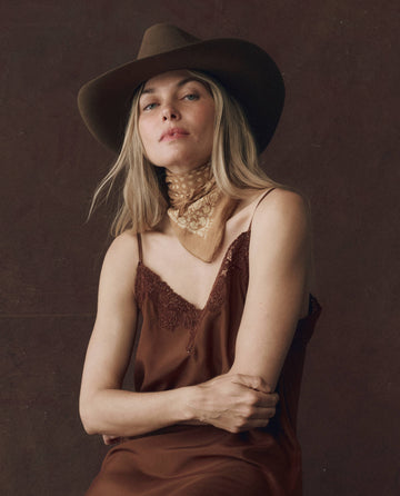 A person wearing a brown hat, The Legacy Dress in Chocolate by THE GREAT. F25 D1 with embroidered mesh lace trim, and a beige neck scarf sits against a dark background.