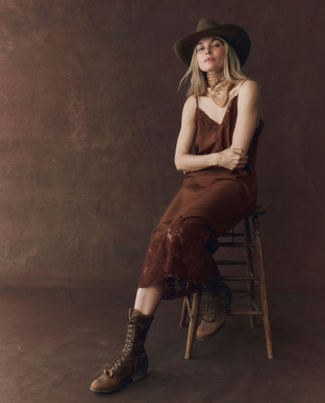 A woman in THE GREAT. F25 D1 The Legacy Dress – Chocolate sits on a wooden stool, wearing brown lace-up boots and a cowboy hat, against a brown textured backdrop.