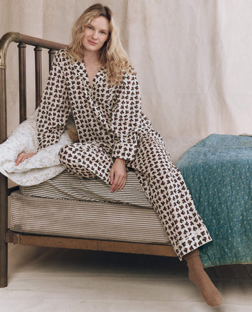 A woman in The Easy Sleep Pant—Cream Horse Medallion Print by THE GREAT. F25 SLEEP sits on a vintage bed with striped bedding and a blue quilt, set against a neutral fabric backdrop.