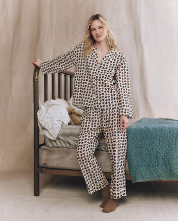 A woman wearing THE GREAT. F25 SLEEP The Easy Sleep Pant in Cream Horse Medallion Print stands by a wooden bed with striped bedding, a blue blanket, and plush toys against a neutral fabric backdrop.