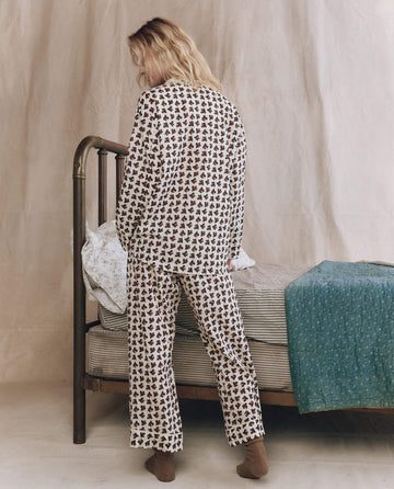 A person wearing The Pajama Shirt in Cream Horse Medallion Print by THE GREAT. F25 SLEEP stands facing away beside a metal-framed bed with striped bedding and a teal blanket in a softly lit room.