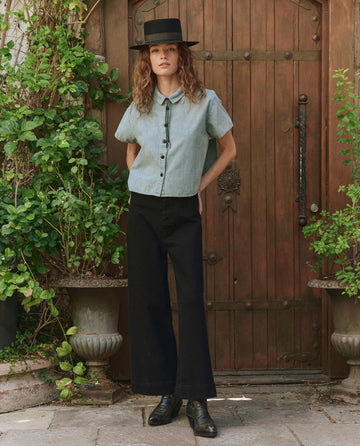 A person wearing THE GREAT. PF25's The Valor Top. -- Boiler Wash, black pants, and boots stands by a wooden door with potted plants, showcasing timeless style.