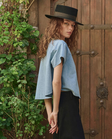 A person models THE GREAT. PF25’s The Valor Top in Boiler Wash—a loose short-sleeve denim shirt—styled with a wide-brimmed black hat and black pants, standing sideways by a wooden door with greenery to the left.