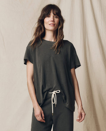 A person with long hair stands against a neutral backdrop, wearing THE GREAT. CORE KNITS' The Boxy Crew in Solid -- Washed Black, paired with drawstring pants. Made from 100% cotton in the U.S.A., this outfit exudes a classic vibe reminiscent of vintage tees.
