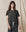 A person with long hair stands against a neutral backdrop, wearing THE GREAT. CORE KNITS' The Boxy Crew in Solid -- Washed Black, paired with drawstring pants. Made from 100% cotton in the U.S.A., this outfit exudes a classic vibe reminiscent of vintage tees.