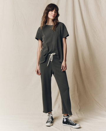 A person wearing a dark casual outfit stands against a neutral backdrop, donning black high-top sneakers and The Boxy Crew. Solid -- Washed Black by THE GREAT. CORE KNITS, made in the U.S.A., paired perfectly with stylish pants.