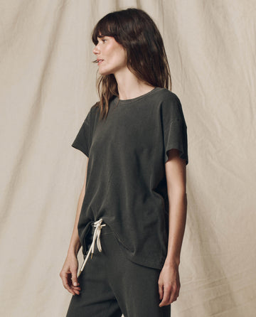 A woman with long hair stands sideways, wearing THE GREAT. CORE KNITS' The Boxy Crew. Solid in Washed Black, a 100% cotton t-shirt, and light-colored drawstring pants. A neutral fabric backdrop complements her timeless U.S.A.-made attire.
