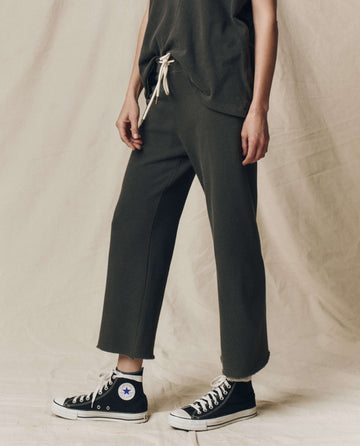 A person in THE GREAT. CORE KNITS' washed black Wide Leg Cropped Sweatpants with a drawstring waist, paired with a matching top and black high-top sneakers with white laces, stands against a beige fabric backdrop.