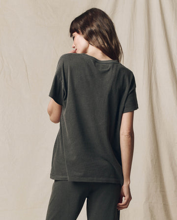 A person with long hair stands facing away, wearing The Boxy Crew in washed black by THE GREAT. CORE KNITS, made from 100% cotton, against a beige fabric background.