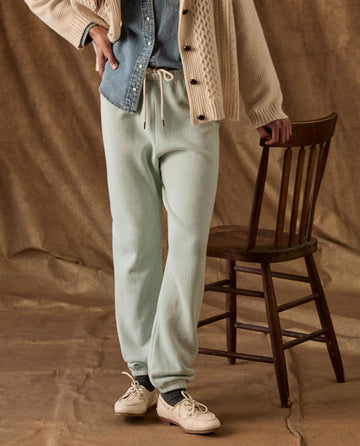 A person wearing The Fleece Stadium Sweatpant in Fresh Mint with a drawstring waist and light-colored loafers, standing next to a wooden chair.