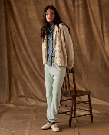 A woman wearing a cream cable-knit cardigan, denim button-down shirt, and The Fleece Stadium Sweatpant in Fresh Mint leans against a wooden chair.