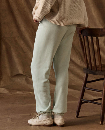 Person wearing vintage mint green The Fleece Stadium Sweatpant with elastic cuffs, a cream sweater, and light-colored loafers, standing next to a wooden chair.