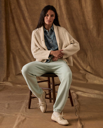 A woman wearing a cream cable-knit cardigan, denim button-down shirt, and The Fleece Stadium Sweatpant in Fresh Mint, sitting on a wooden stool.