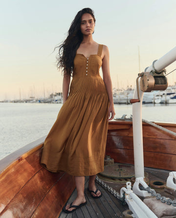 A woman in THE GREAT. SU26's The Day Break Dress in Clay and black sandals stands on a wooden boat, with a marina and docked sailboats in the background.