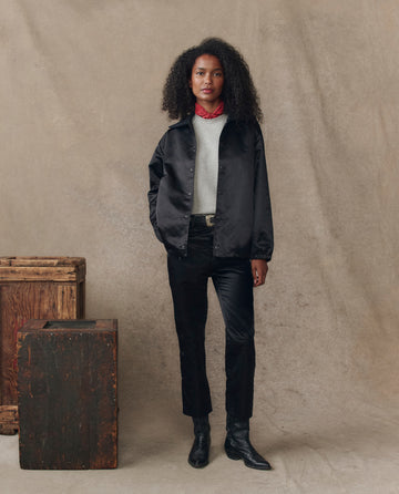 A woman stands indoors by two wooden crates, wearing THE GREAT. H25 COLLECTION’s The Satin Coach's Jacket in black over a white top, black pants, black boots, and a red necklace against a neutral backdrop.