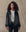 A woman faces forward against a beige background wearing THE GREAT. H25 COLLECTION's The Satin Coach's Jacket in black over a grey shirt, paired with black pants and a red bandana around her neck.