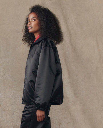 A person with curly hair wears THE GREAT. H25 COLLECTION The Satin Coach's Jacket in black, paired with matching pants, standing against a neutral beige background.