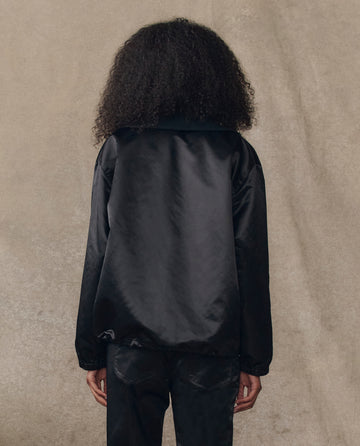 A person with curly hair, seen from behind, wears THE GREAT. H25 COLLECTION’s The Satin Coach's Jacket in black, paired with black pants, in front of a neutral textured background.