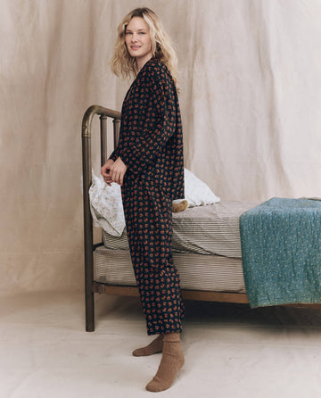 A woman stands beside a metal-framed bed, wearing THE GREAT. F25 SLEEP The Easy Sleep Pant in Navy Horse Medallion Print and brown socks, with bedding and a blue blanket on the bed.