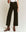 A person wearing THE GREAT. F25 D1 The Seafair Jean.. -- Carbon, a white top, suspenders, and brown ankle boots stands against a plain background.