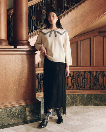 A person wearing The Bow Sweater. in cream with a grey neckline detail and a black fringe skirt, showcasing women’s fashion.