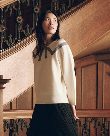 A person wearing The Bow Sweater in Cream, featuring a grey chevron pattern across the chest and shoulders, paired with a black skirt.