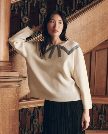 A woman wears The Bow Sweater in cream, featuring a black graphic across the neckline, paired with a black skirt.