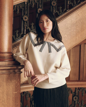 A woman wearing The Bow Sweater in cream, a knit sweater with a V-shaped neckline accented by a dark, textured bow.