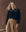 A woman with long blonde hair wears THE GREAT. F25 D1 The Crochet Collar Pullover in black with soft white, styled with a tan pleated skirt, standing before a brown background.