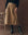 A person wearing THE GREAT. F25 D1 The Field Skirt in Tobacco, paired with a black sweater, black belt, and black cowboy boots stands next to a wooden chair against a brown background.