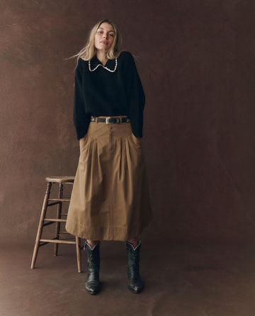 A woman stands beside a wooden stool against a brown backdrop, wearing THE GREAT. F25 D1's Field Skirt in Tobacco, a black sweater with a white collar, a black belt, and black cowboy boots.