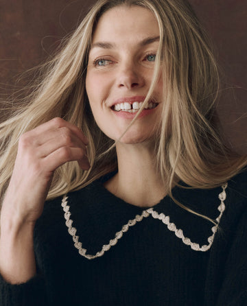 A woman with long blonde hair smiles and looks to the side, wearing THE GREAT. F25 D1's The Crochet Collar Pullover in black with soft white.