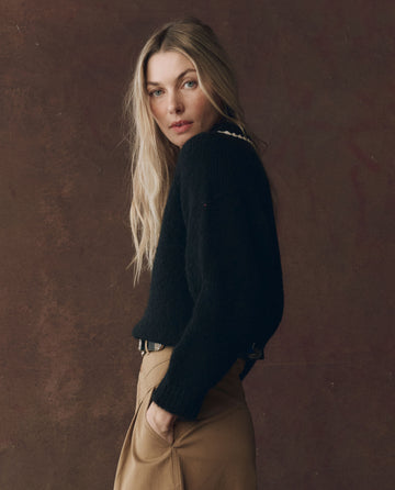 A woman with long blonde hair models THE GREAT.’s Crochet Collar Pullover in Black with Soft White, paired with tan pants, standing against a plain brown background and glancing over her shoulder.