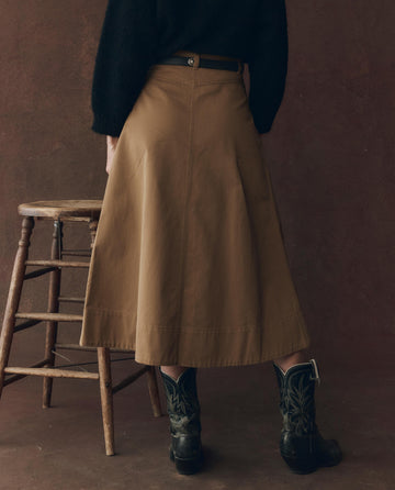 A person stands next to a wooden stool, seen from behind, wearing a black top, black cowboy boots, and THE GREAT. F25 D1 The Field Skirt in Tobacco.